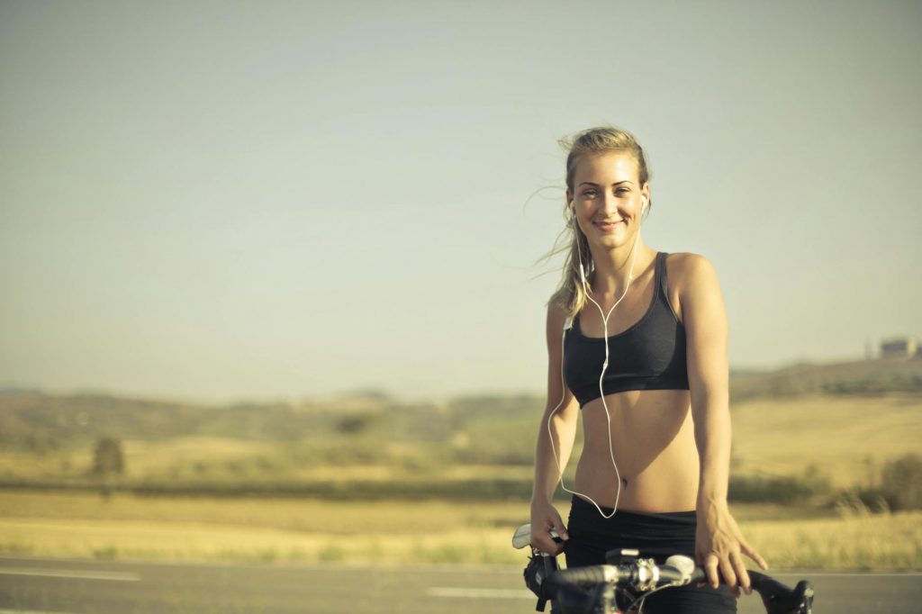 selective focus photo of smiling woman in black active wear with earphones on standing next to her bicycle by the side of the road