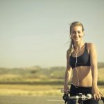 selective focus photo of smiling woman in black active wear with earphones on standing next to her bicycle by the side of the road