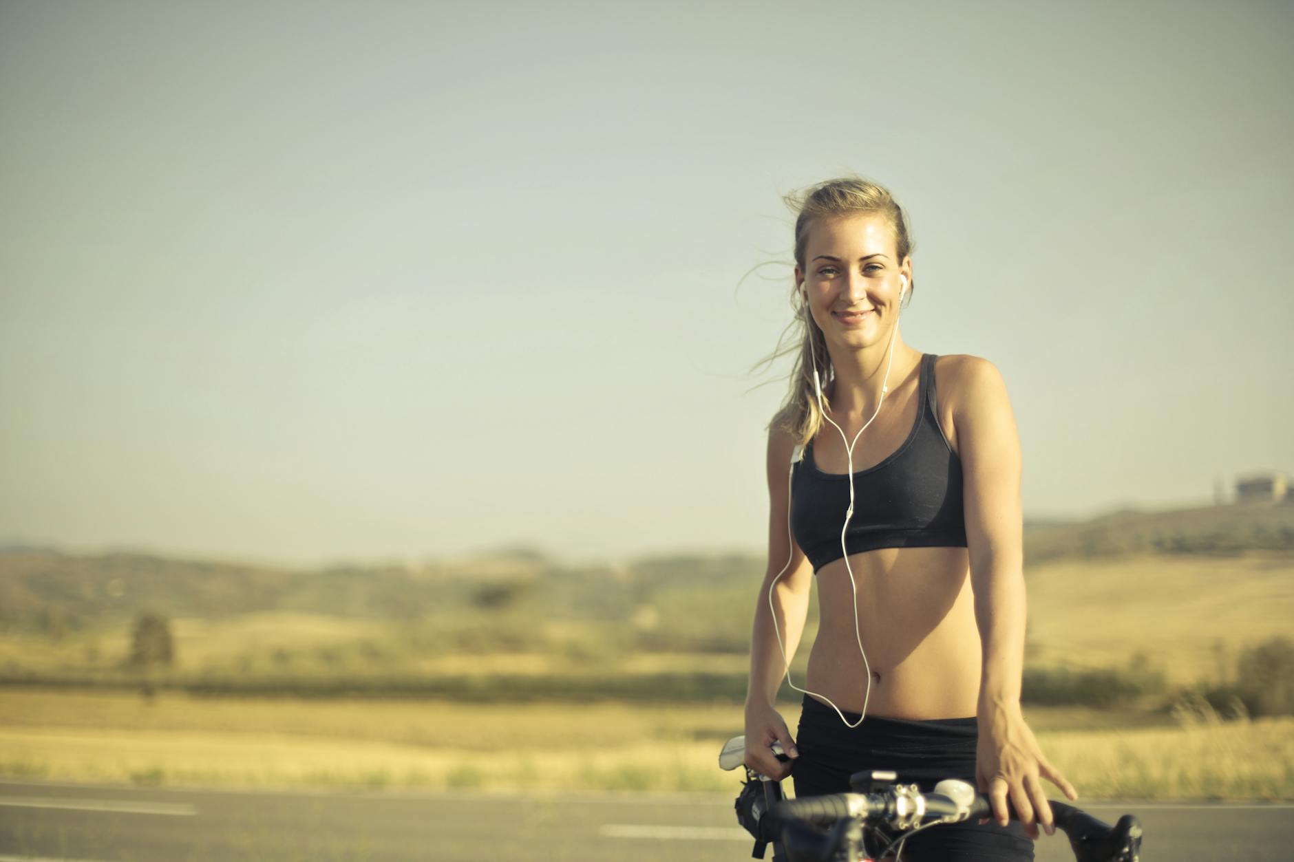 selective focus photo of smiling woman in black active wear with earphones on standing next to her bicycle by the side of the road